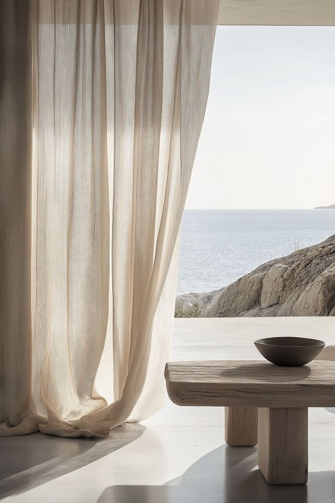 Sunlight through soft linen curtains beside an open sea view and a pale wooden table.
